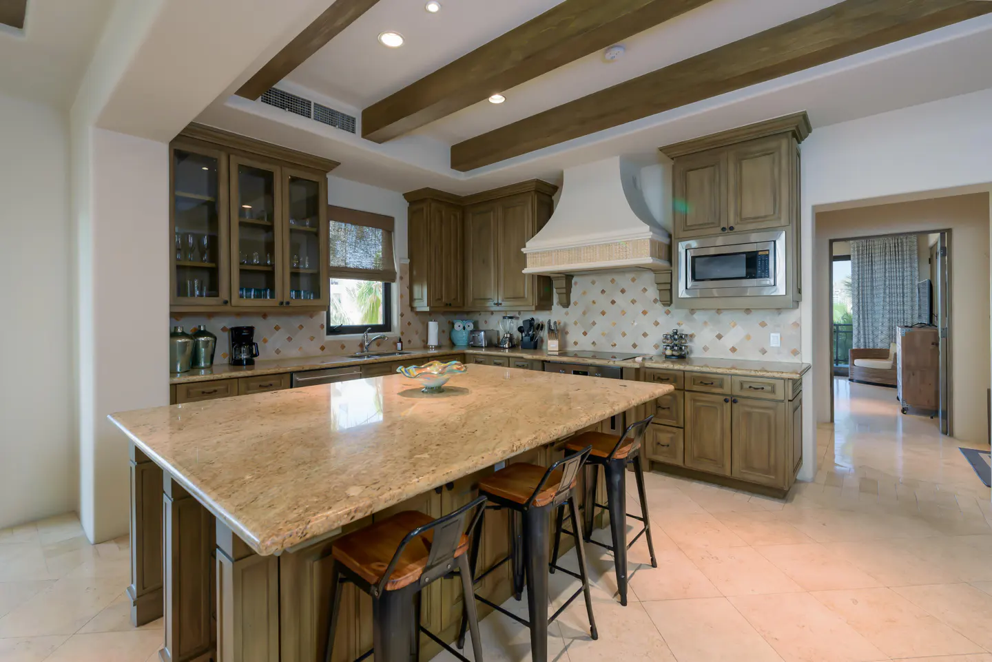 A kitchen with tan cabinets, a large island with stools, and wooden beams on the ceiling.