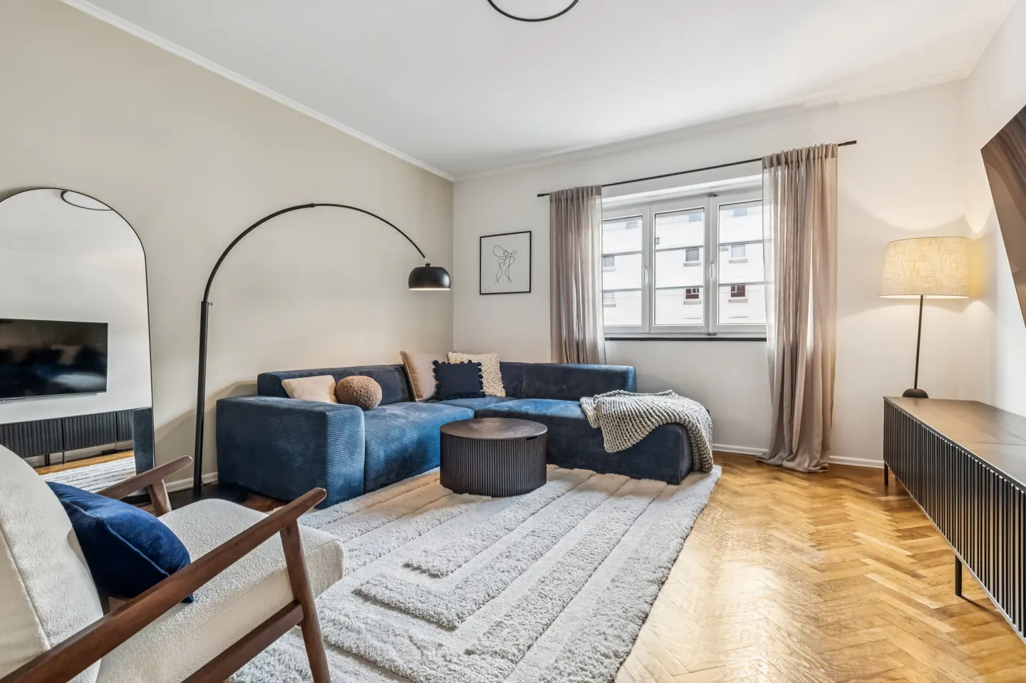 Living room with blue sectional sofa, round coffee table, patterned rug, and herringbone wood floors. A black arc lamp and a window with curtains are visible.