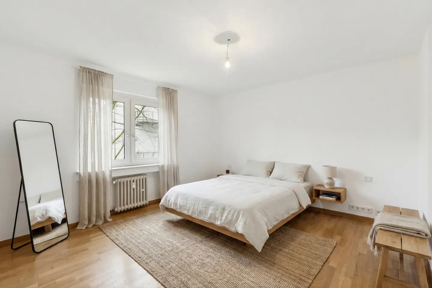 Bright bedroom with white walls, wooden floor, and a jute rug. A bed with white linens, a black framed mirror, and a window with beige curtains are visible.