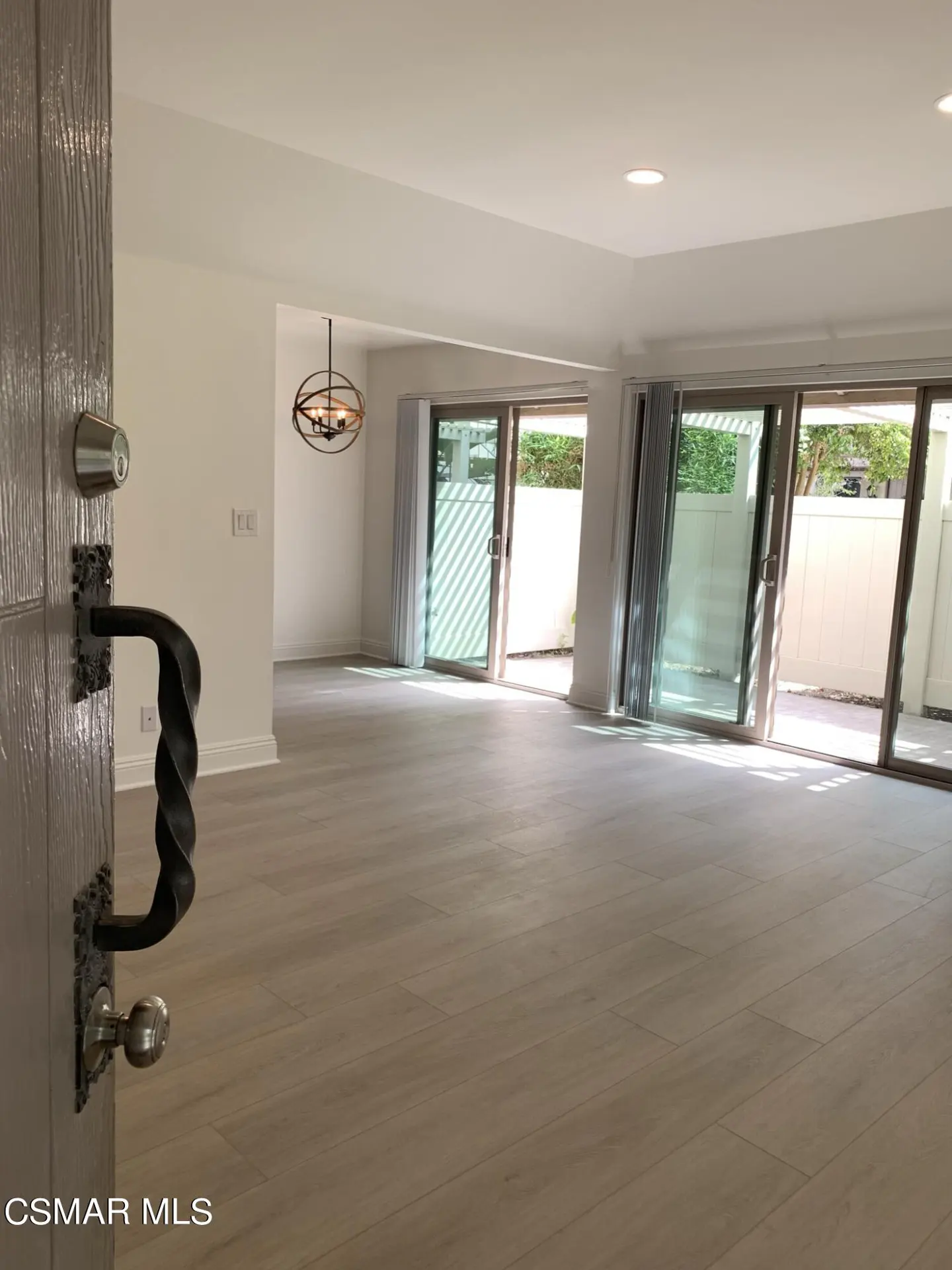 Open front door reveals a bright, empty living room with light wood floors and sliding glass doors to a patio. A modern chandelier hangs in the background.