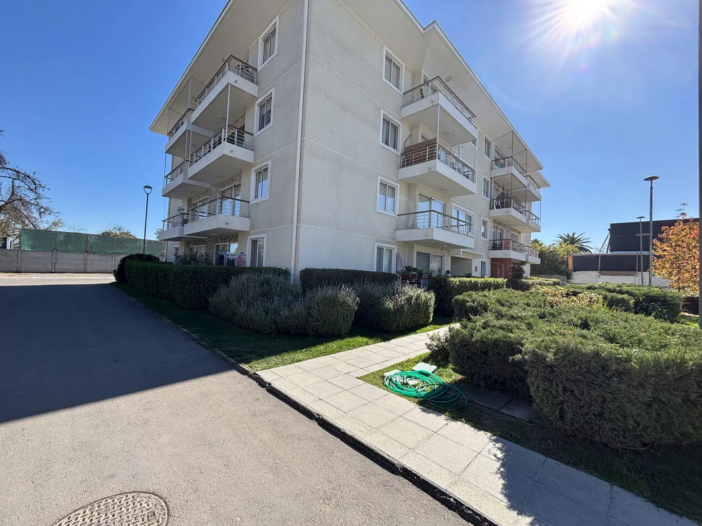 Exterior view of a four-story apartment building with white balconies, surrounded by green bushes and a sidewalk.