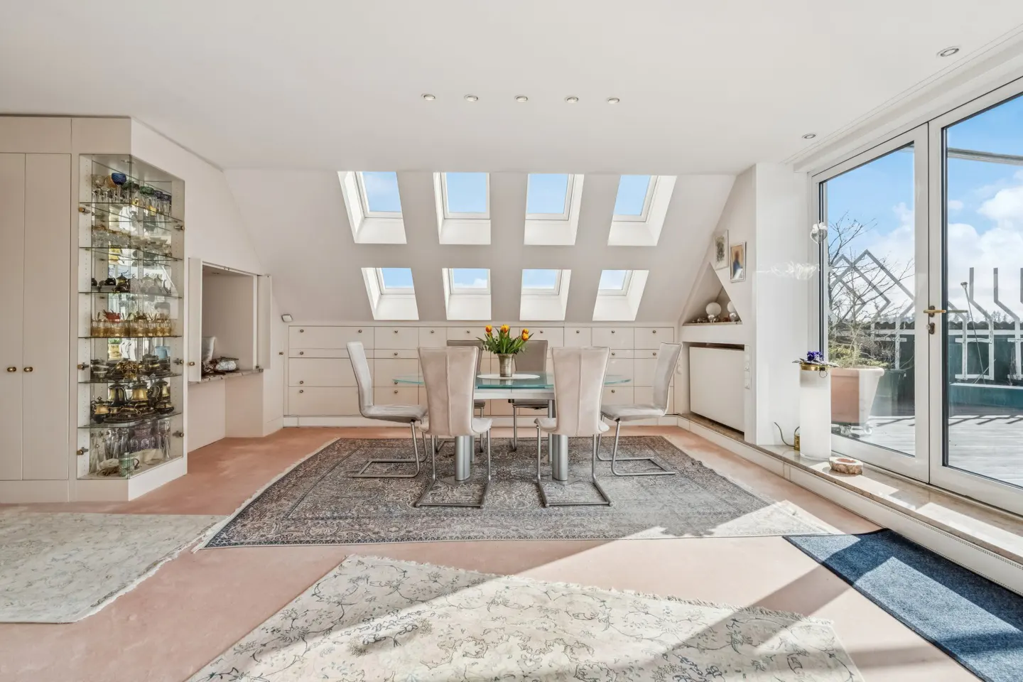 Bright dining room with skylights, glass table, and beige chairs on a patterned rug. Balcony access with a view.