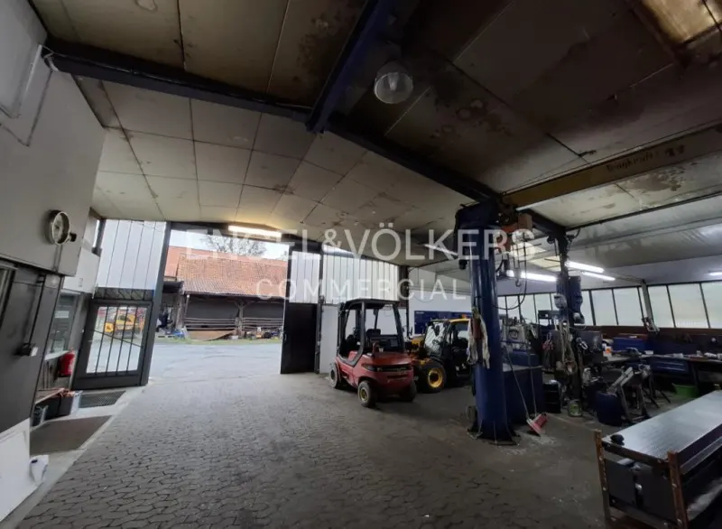 Interior view of a commercial garage with a forklift, tractor, and car lift. The space has an open door and a concrete floor.