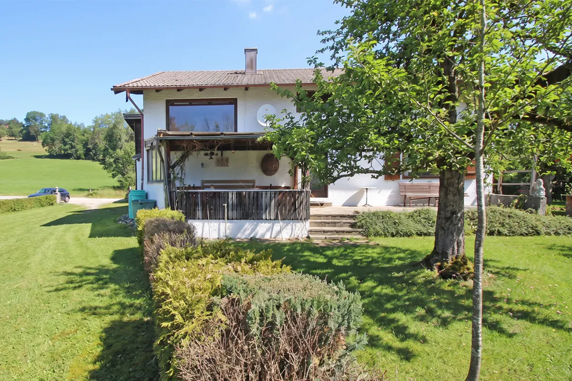 Exterior view of a white two-story house with a brown roof, a porch, and a green lawn. A large tree stands in the yard.