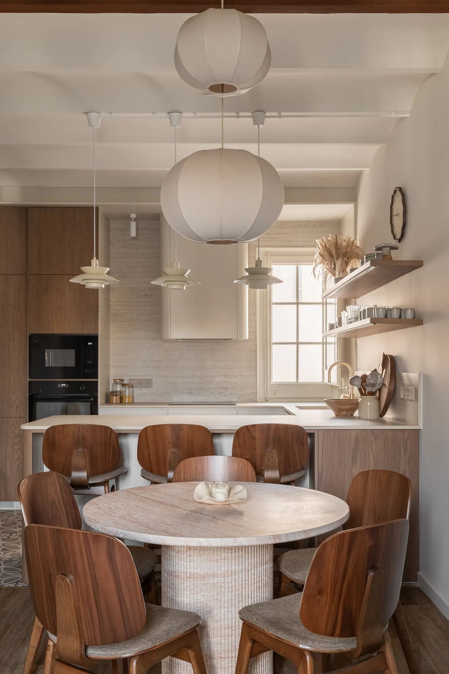 A bright, modern kitchen features a round marble table with six wooden chairs, pendant lights, and wooden cabinets.