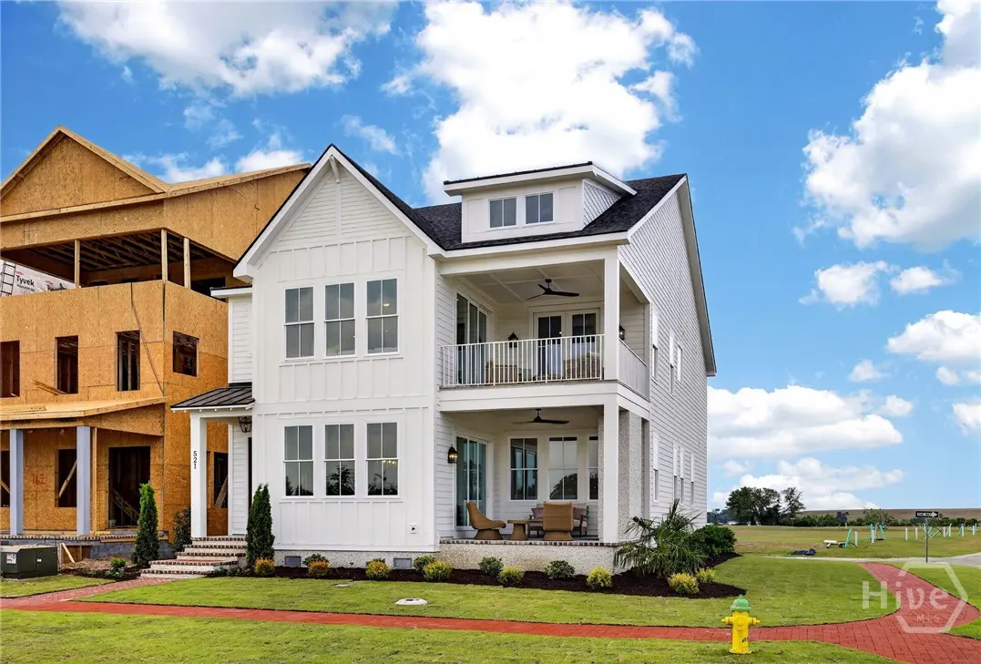 Two-story white house with balconies, black roof, and green lawn under a blue sky. A house under construction is next door.