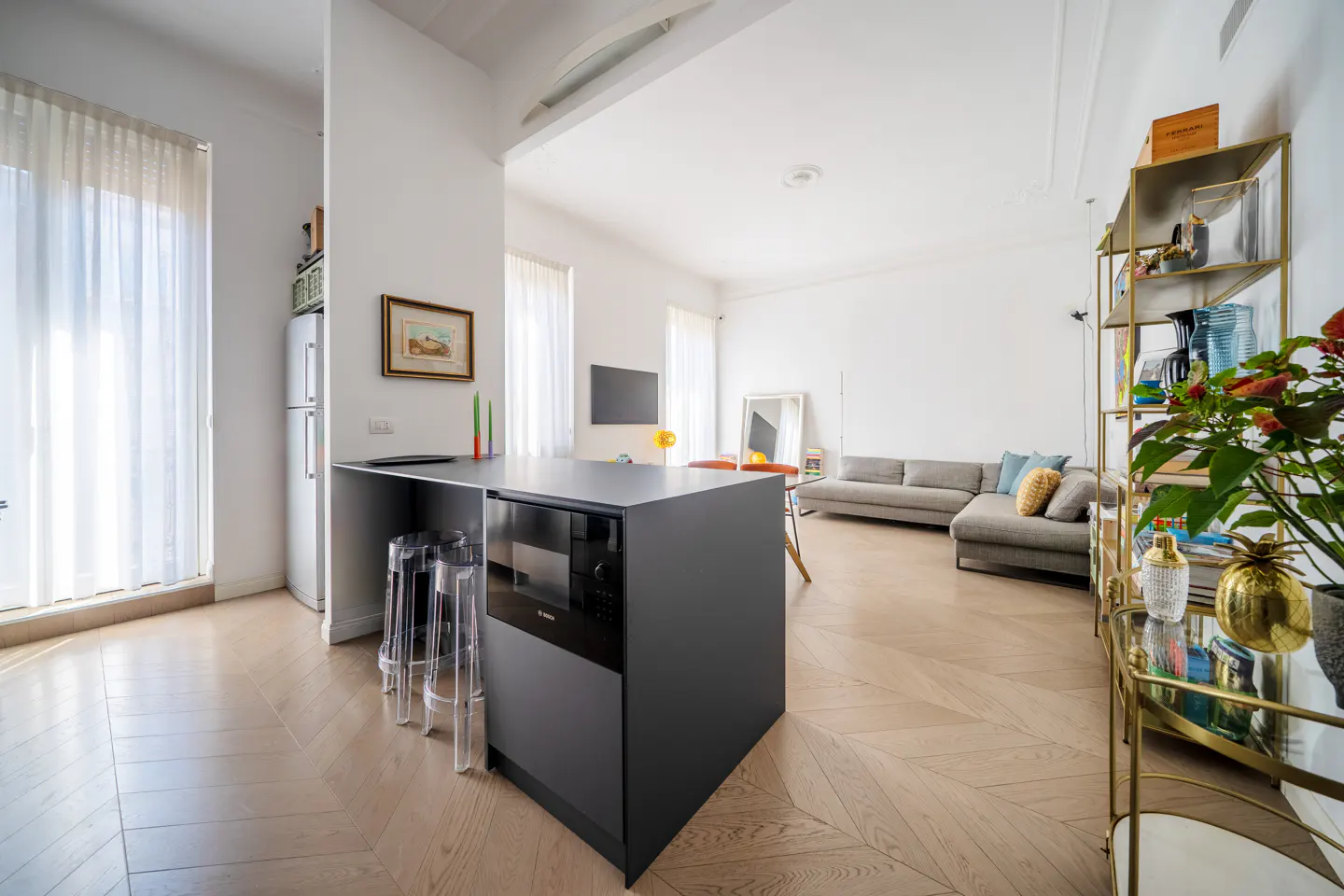 Bright, open-concept living space with a gray kitchen island, clear stools, and herringbone wood floors. A gray sofa and gold shelving unit are in the background.