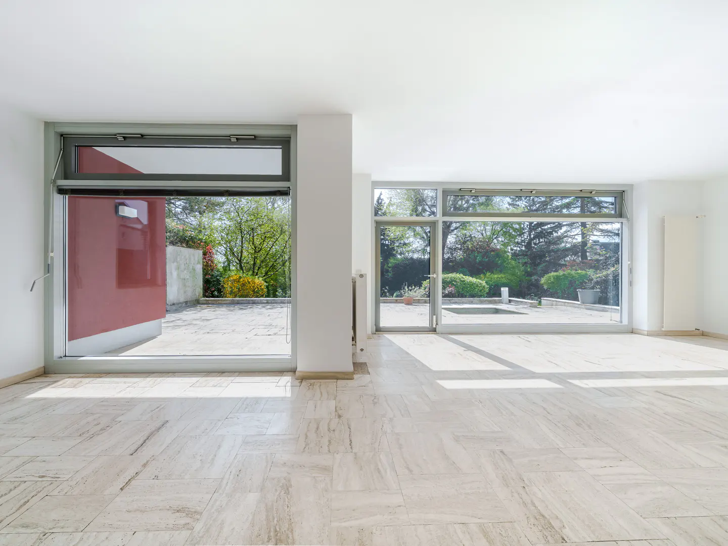 Bright, empty room with marble floors and large windows. Outside, a patio and garden are visible. A red door is partially open.