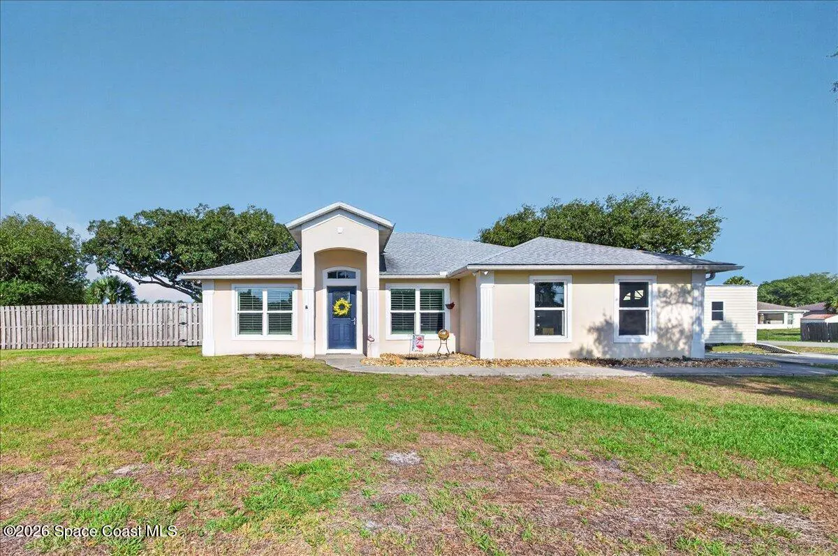 Beige single-story house with a blue door and a sunflower wreath, green lawn, and a blue sky.