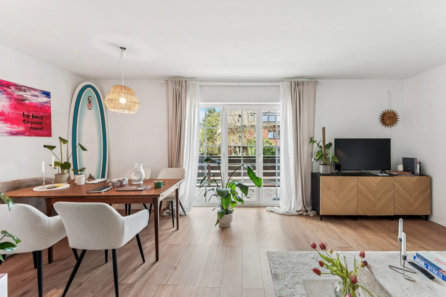Bright living room with wood floors, a dining table with white chairs, and a TV on a wooden cabinet. Balcony doors with sheer curtains let in natural light.