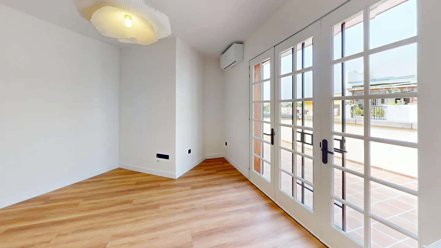 Bright room with wood floors, white walls, and a modern light fixture. French doors open to a patio with a view of a nearby building.