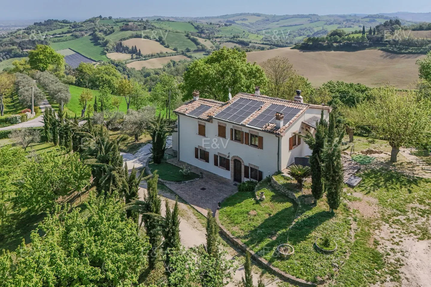 Aerial view of a cream-colored house with brown shutters and solar panels, surrounded by lush green trees and rolling hills.