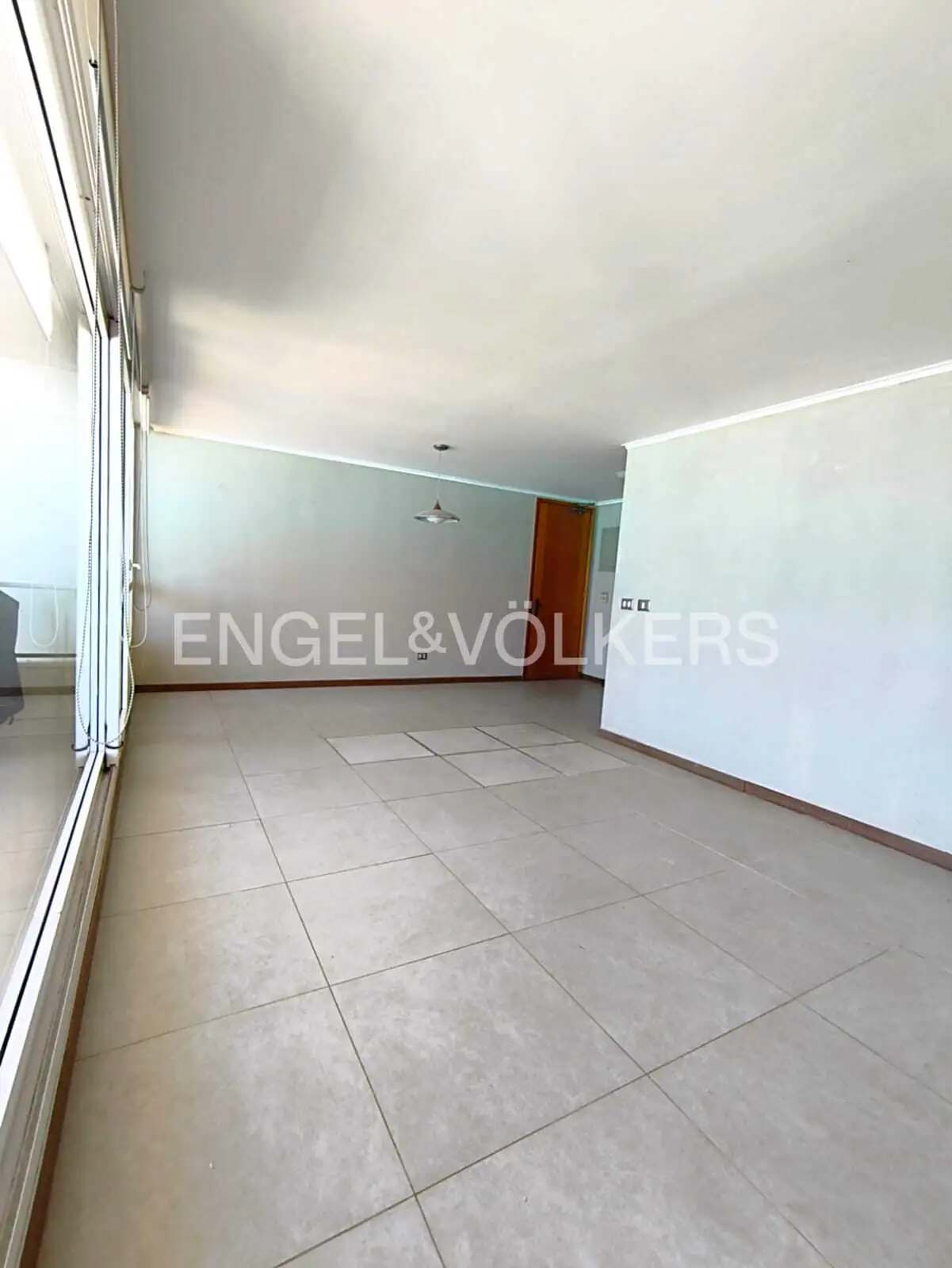 Spacious, empty room with light gray tiled floors, white walls, large windows on the left, and a wooden door in the back.