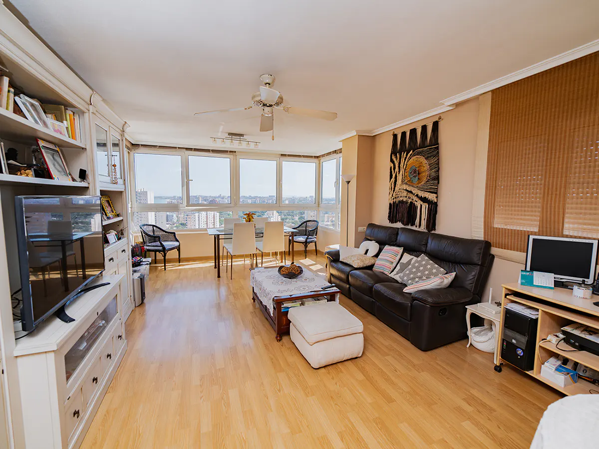 Bright living room with wood floors, a black leather sofa, and a large window overlooking a city. A white bookshelf and TV stand are on the left.