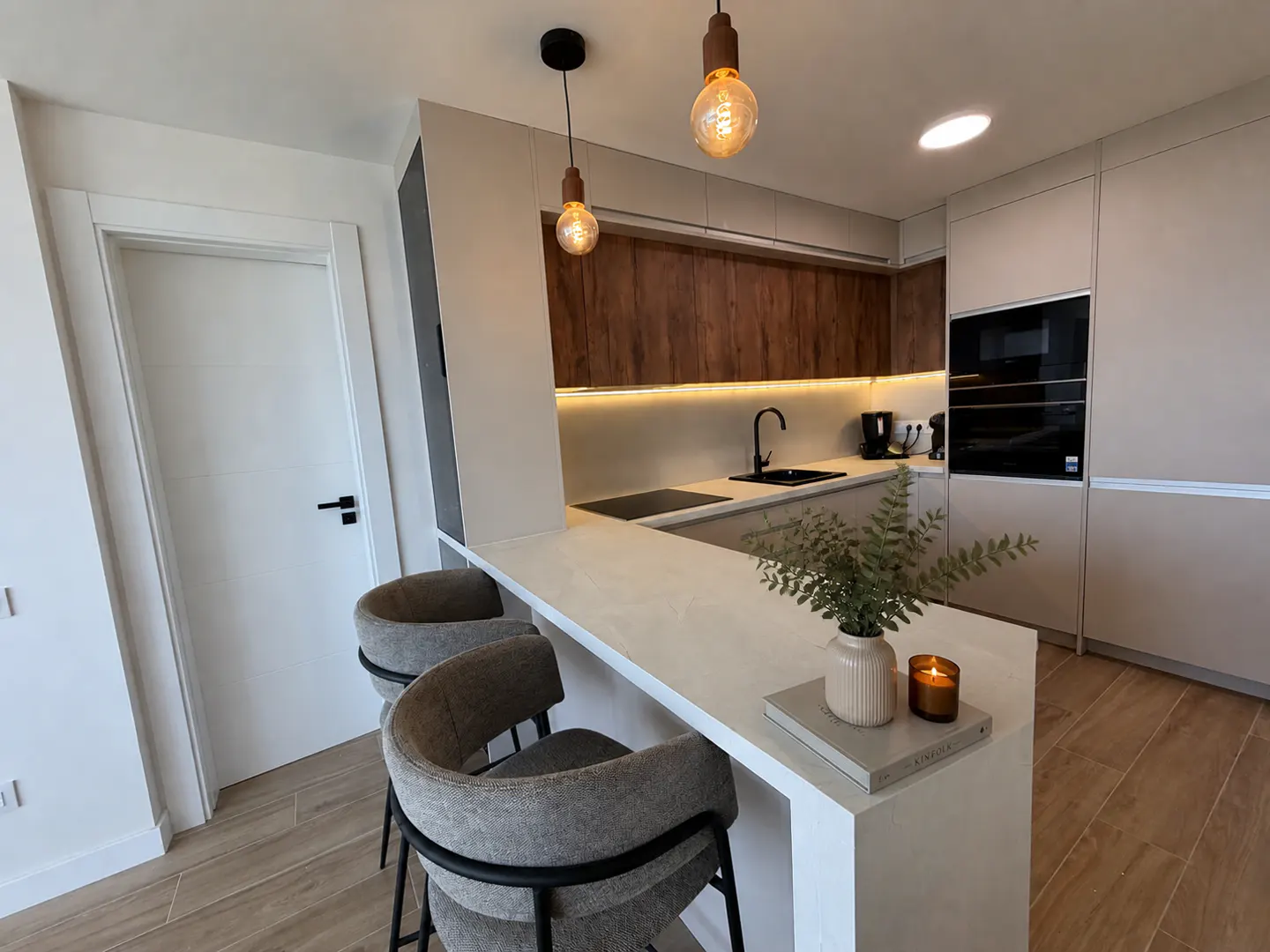 A modern kitchen with a white countertop island, two gray bar stools, and wood cabinets. Pendant lights hang above the island.