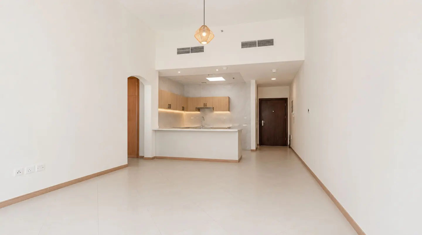 Bright, empty apartment with white walls and tile floors. A kitchen with light wood cabinets and a white island is visible. A brown door is on the right.