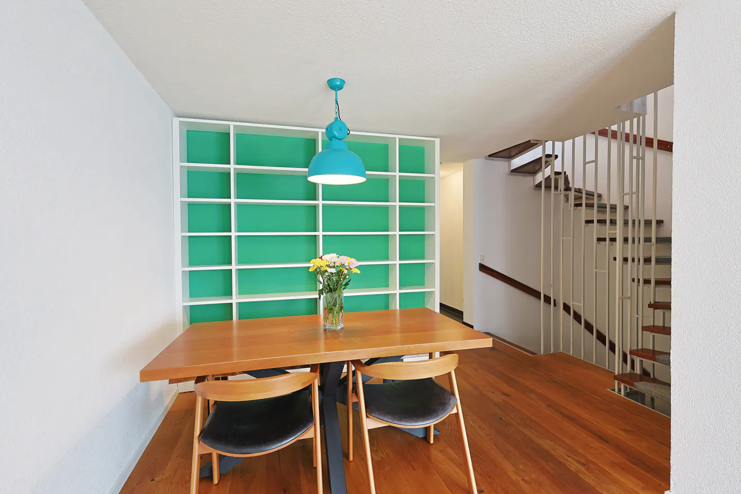 Dining area with wooden table, black chairs, a bright teal pendant light, and a green bookshelf against the wall.