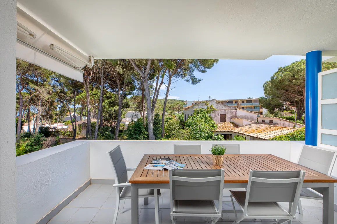 Covered balcony with a wooden dining table, grey chairs, and a potted plant, overlooking trees and Mediterranean houses.