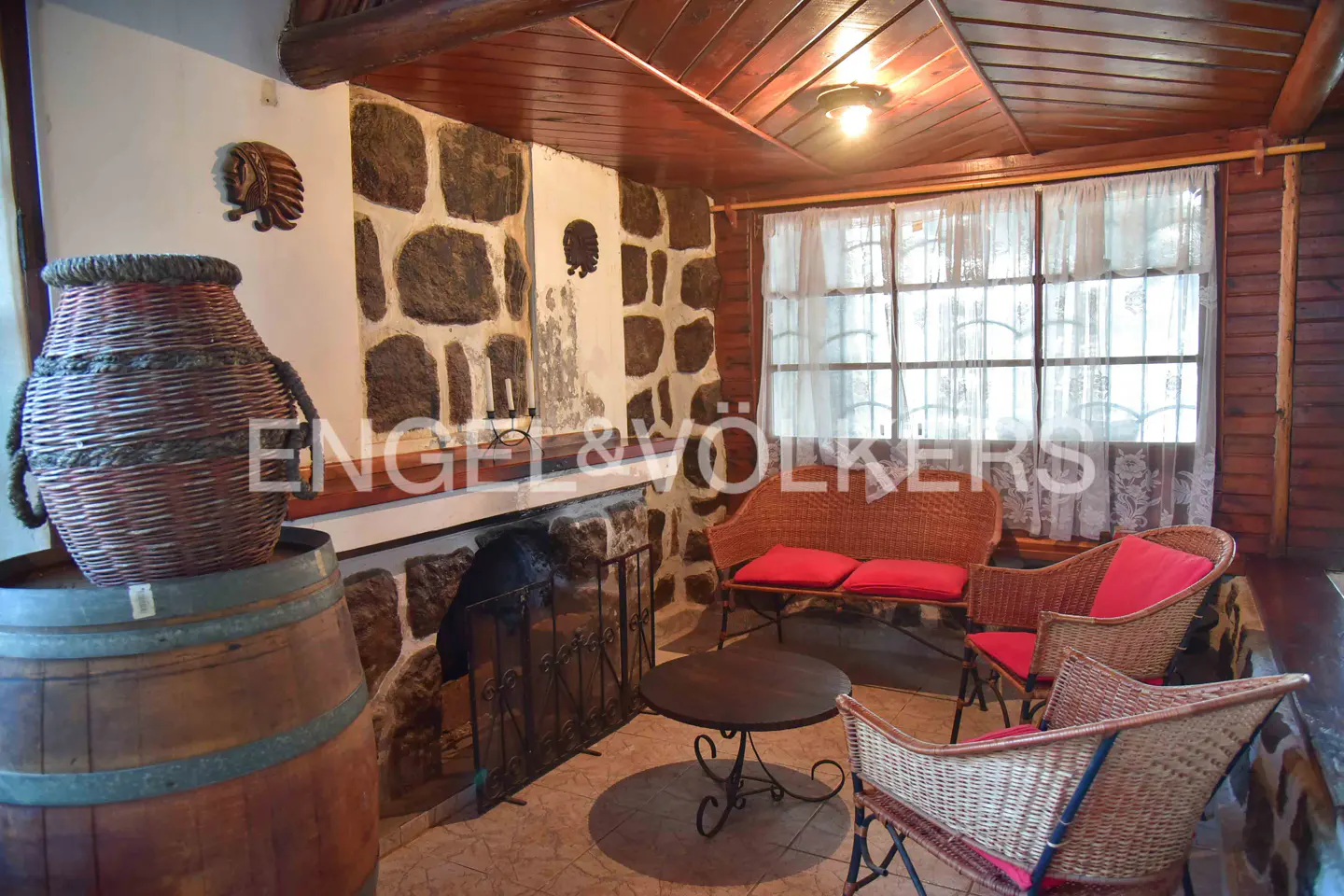Rustic sunroom with stone fireplace, wicker furniture with red cushions, and a wooden barrel topped with a woven basket.