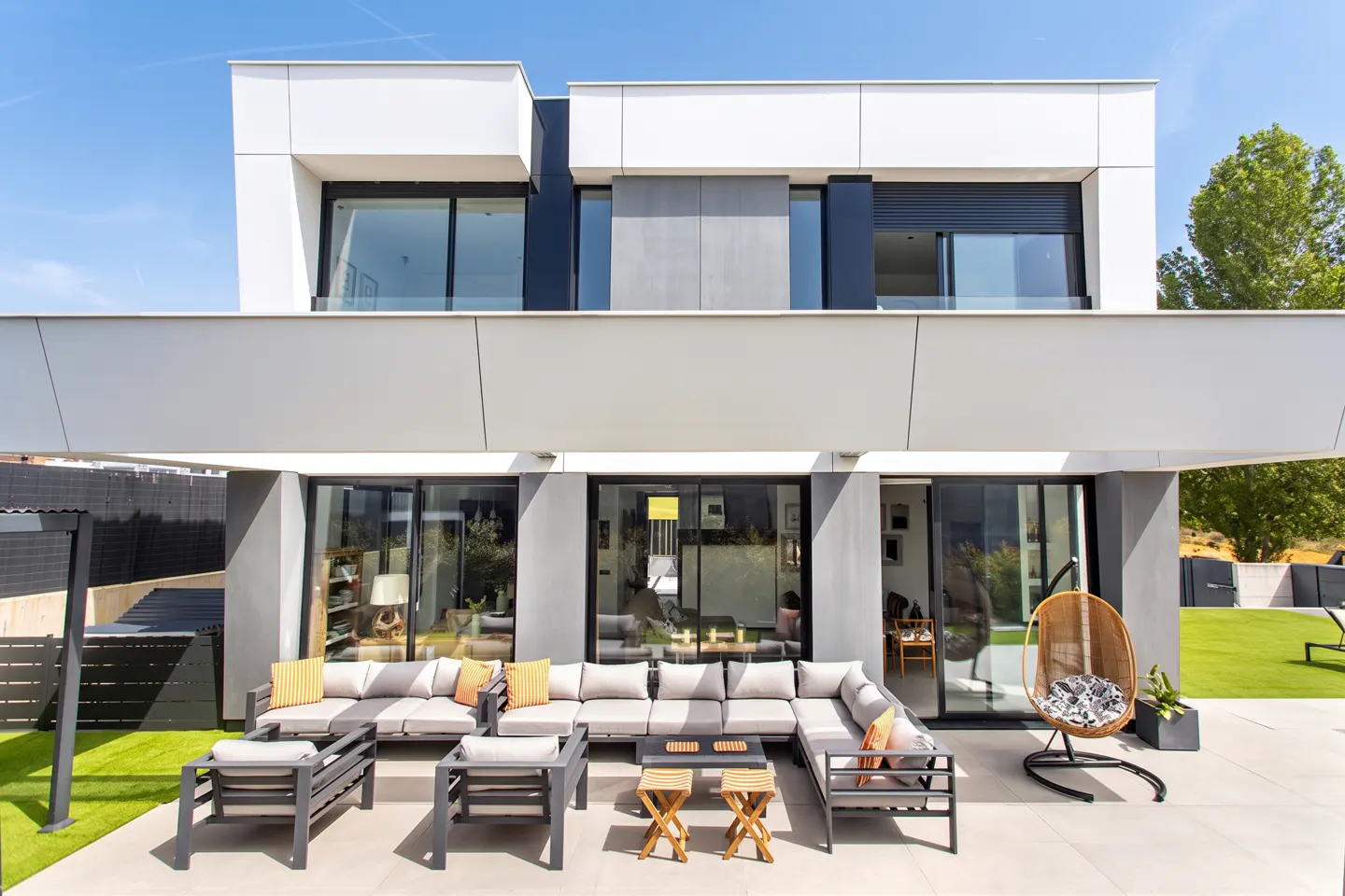 Modern two-story house with a patio featuring gray outdoor furniture and a hanging chair on a sunny day.