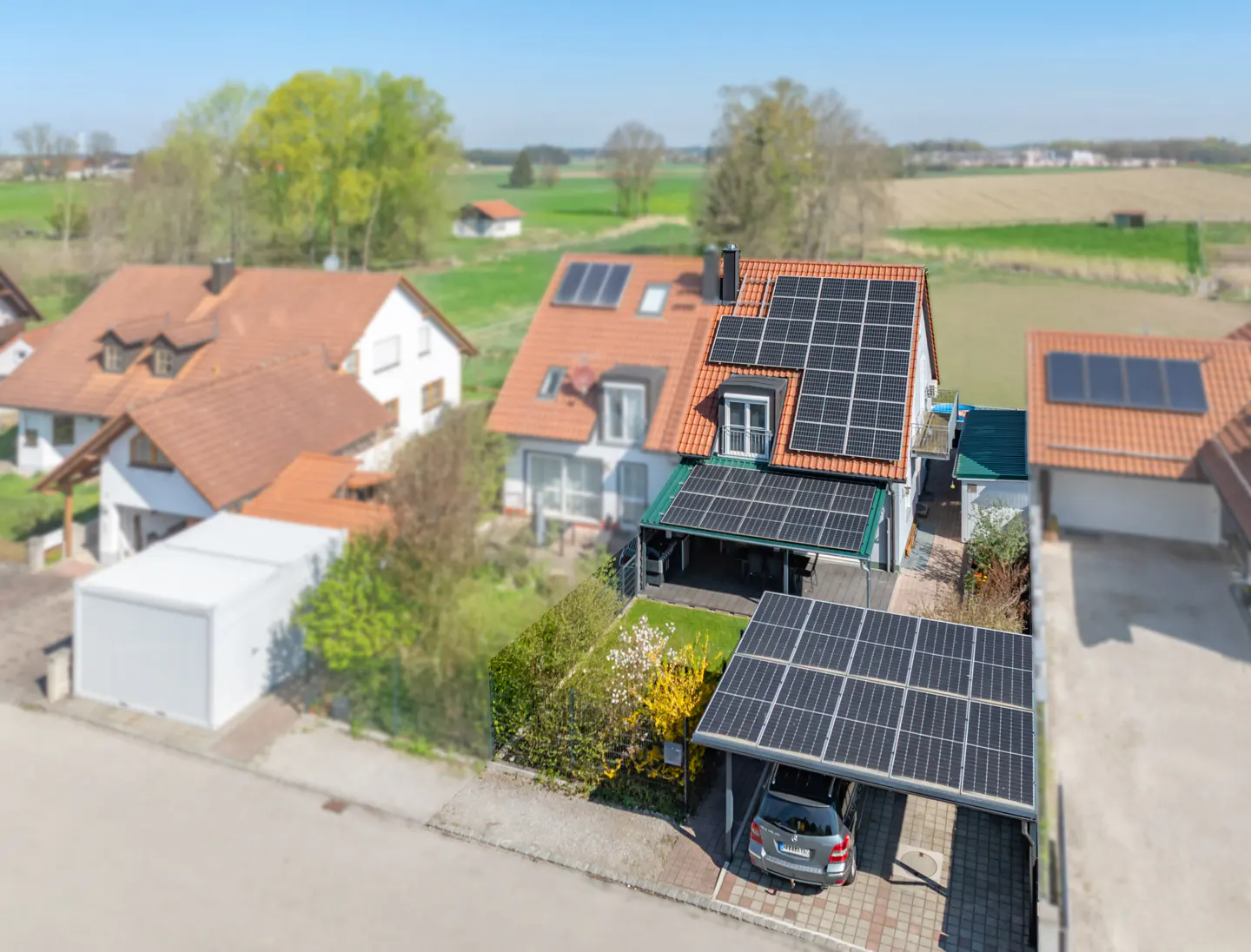Aerial view of a house with solar panels on the roof and carport, surrounded by green fields and other houses.