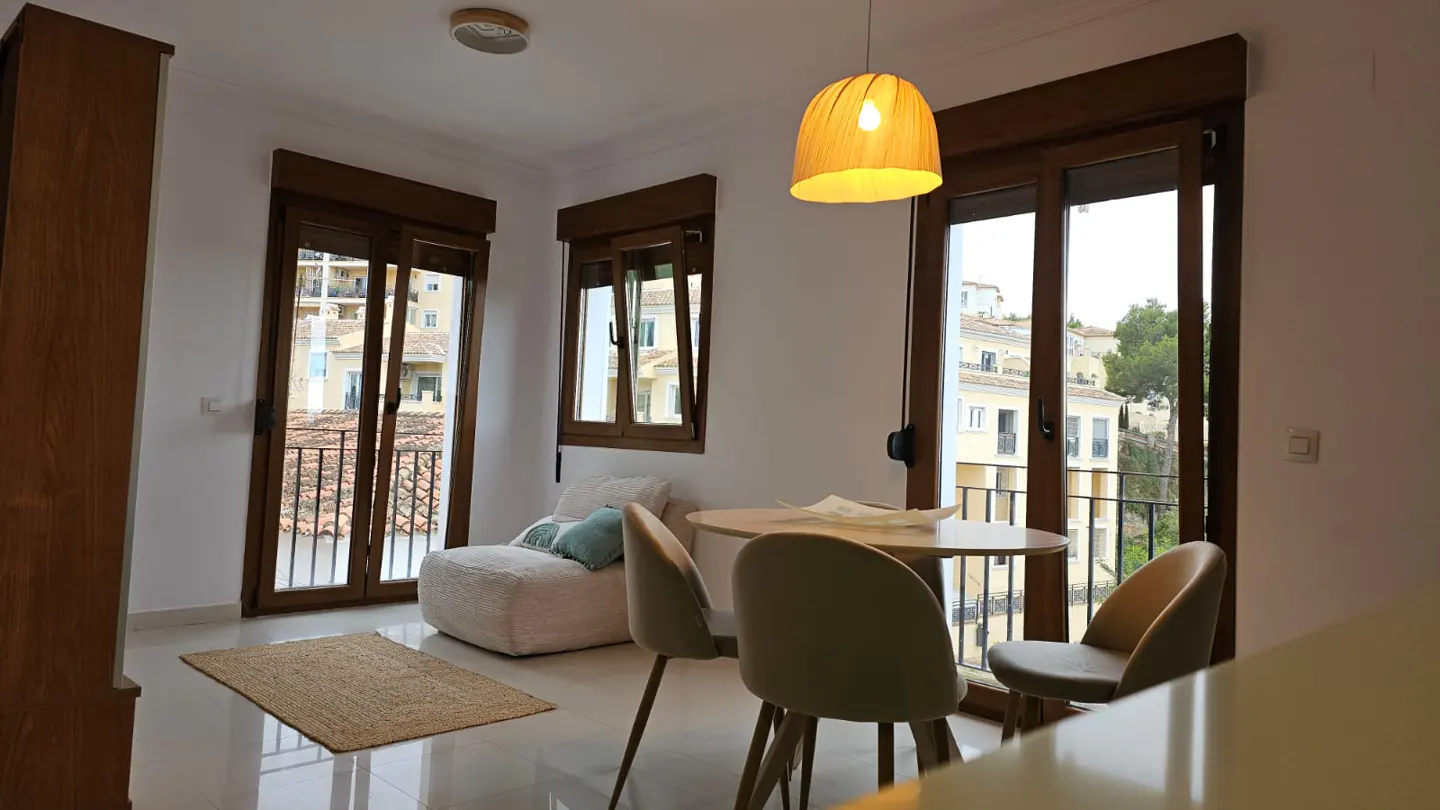 Bright room with a round table, chairs, and a fluffy chair near a window with a view of buildings. A straw pendant lamp hangs above.