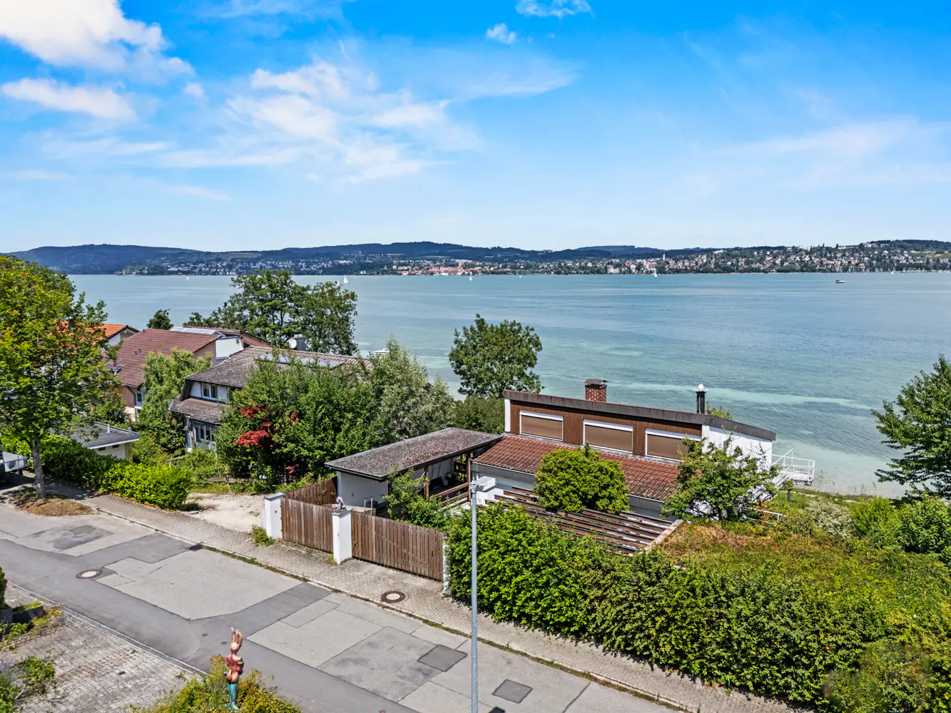 Scenic view of houses near a lake under a blue sky with white clouds. Lush green trees and bushes surround the properties.