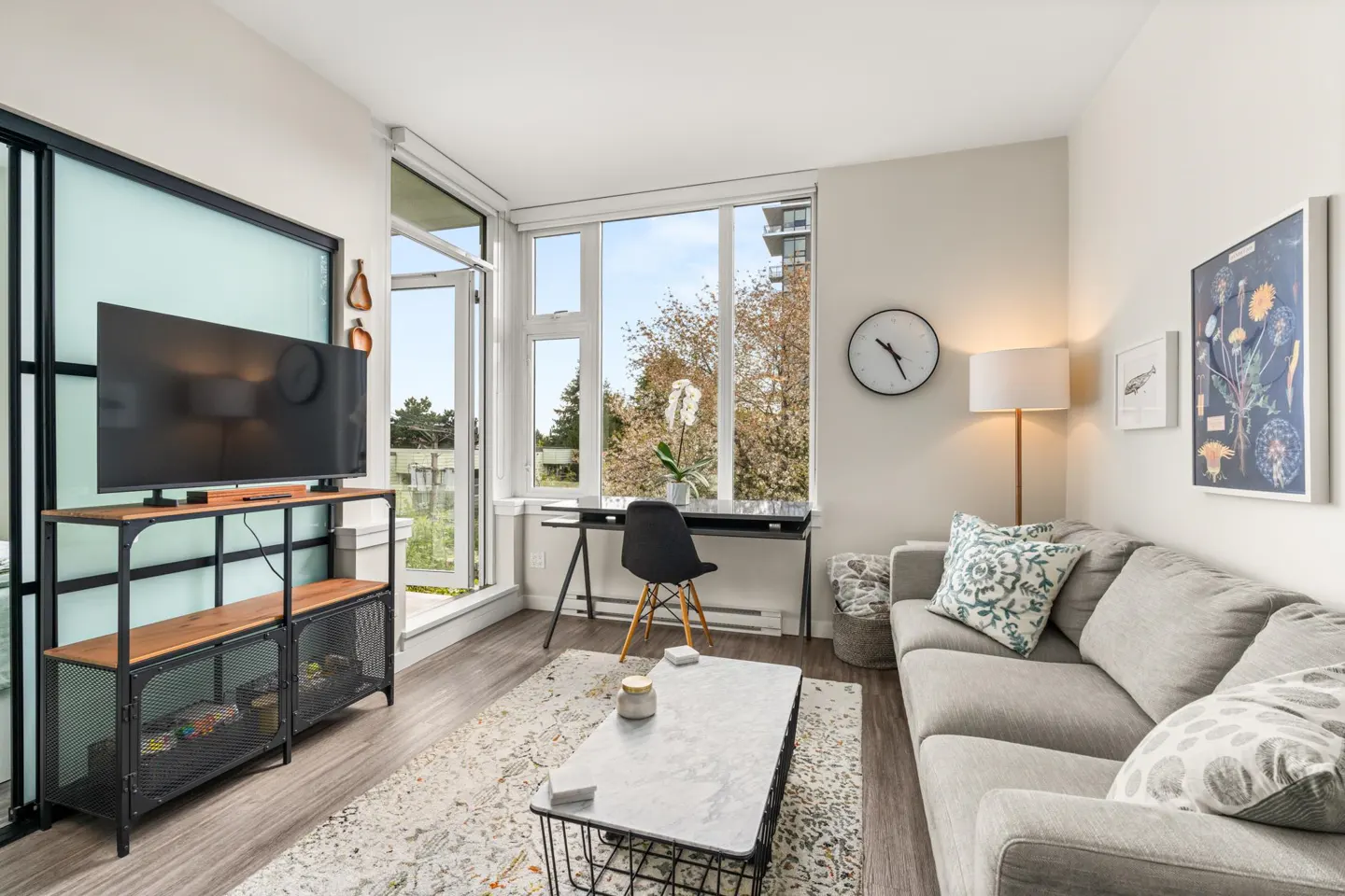Bright living room with a gray sofa, patterned pillows, a black desk by a window, and a TV on a wooden stand.