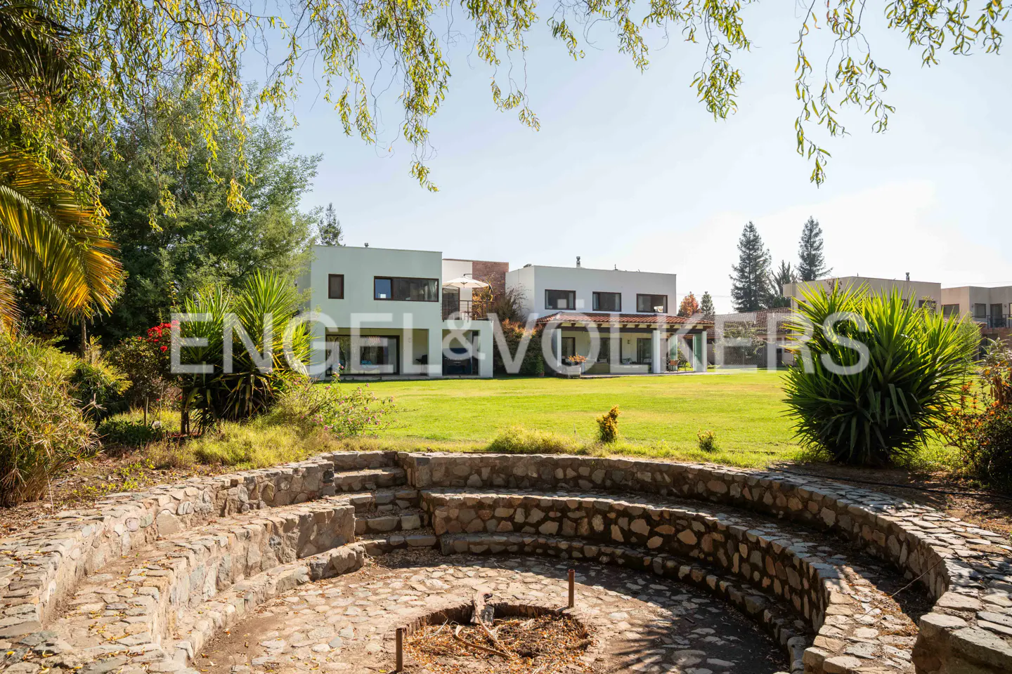 Stone fire pit in front of modern white houses with green lawn and trees under a blue sky.