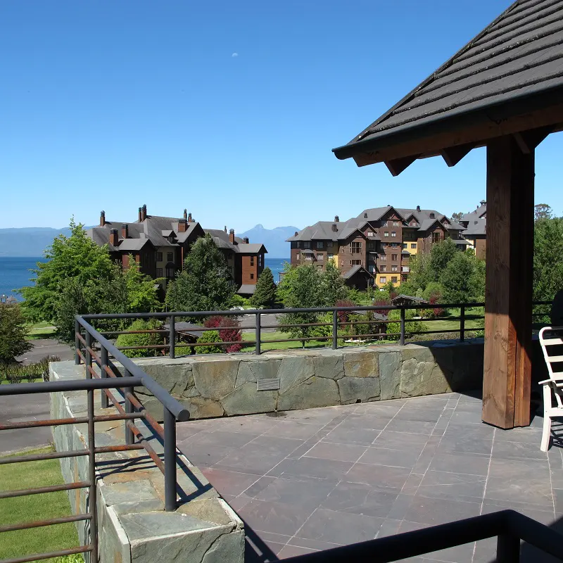 View from a stone patio with a gazebo overlooking brown buildings, green trees, and a blue lake under a clear sky.