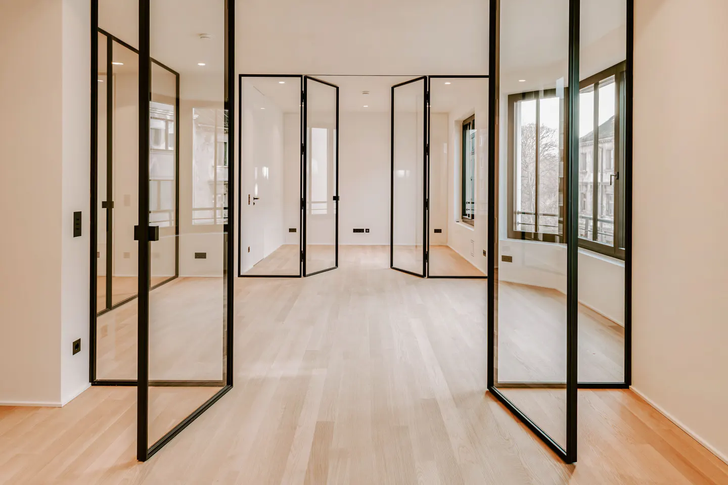 Bright room with light wood floors and white walls. Black framed glass doors are partially open, leading to another room with windows.