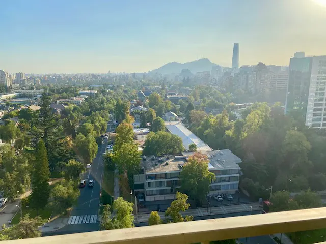 City view from a balcony. Buildings and trees stretch to the horizon under a blue sky. A tall skyscraper is visible in the distance.