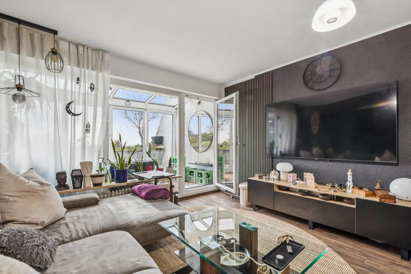 Modern living room with a grey sectional sofa, glass coffee table, large wall-mounted TV, and a bright sunroom entrance.