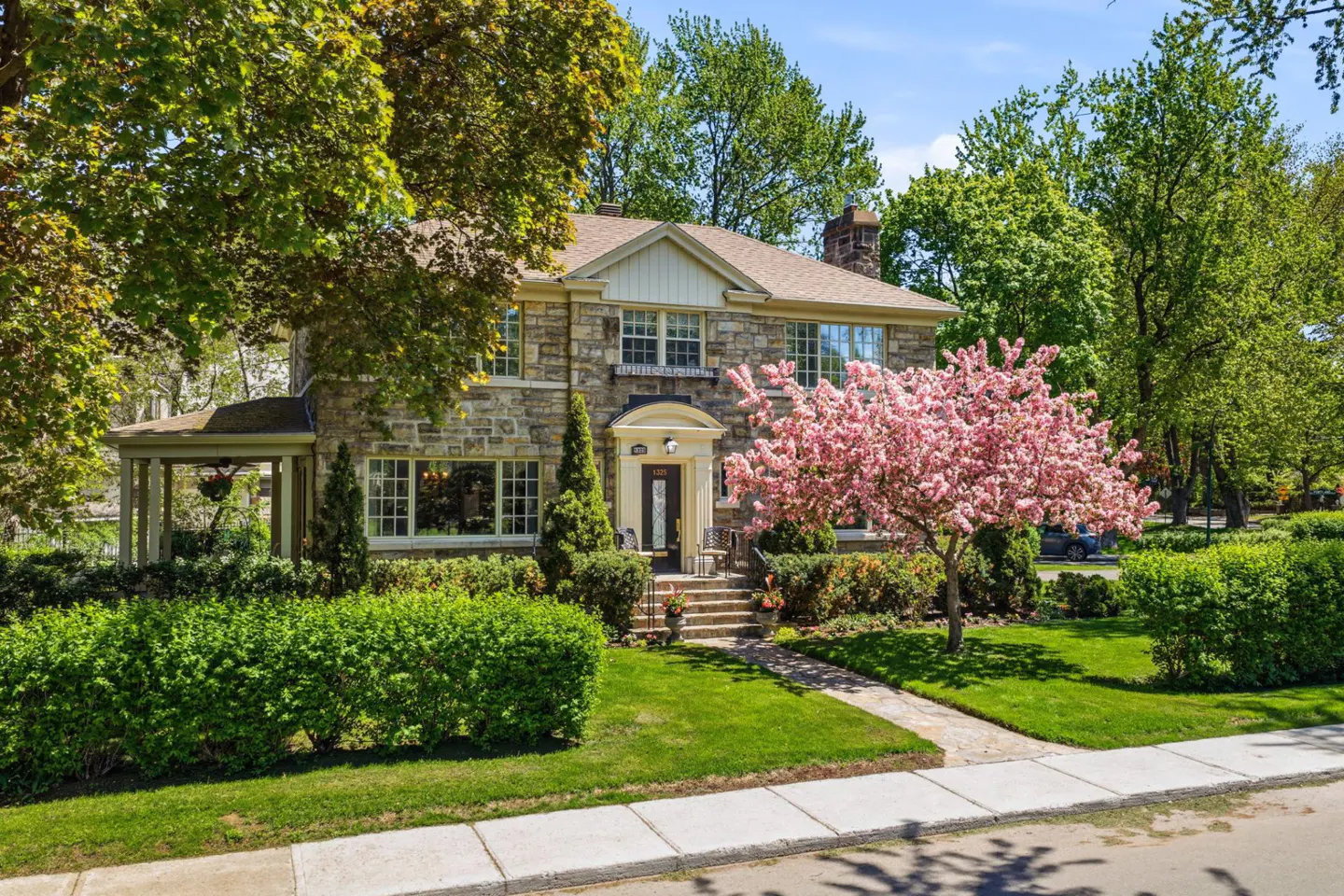 Two-story stone house with a brown roof, green lawn, and a pink flowering tree in the front yard.