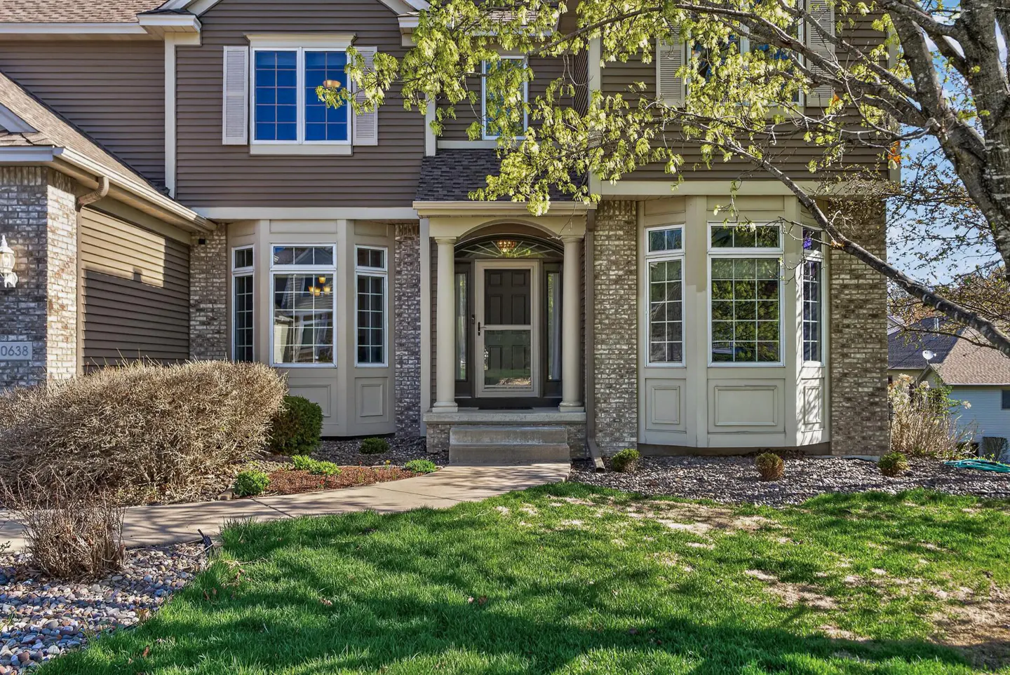 Two-story house with brown siding, brick accents, and bay windows. Green lawn and walkway lead to the front door.