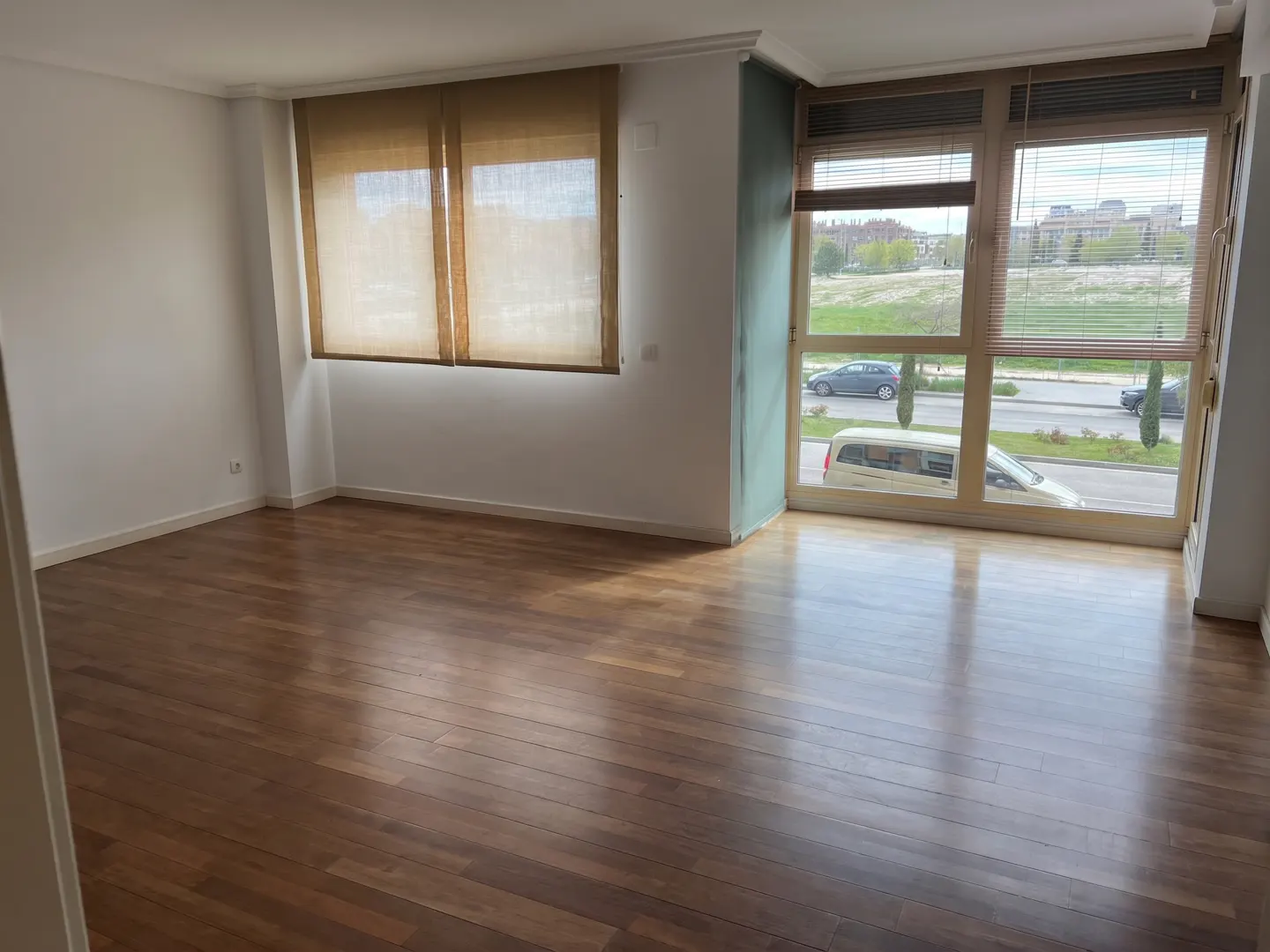 Empty room with dark wood floors, white walls, tan window shades, and large windows overlooking a street and green field.