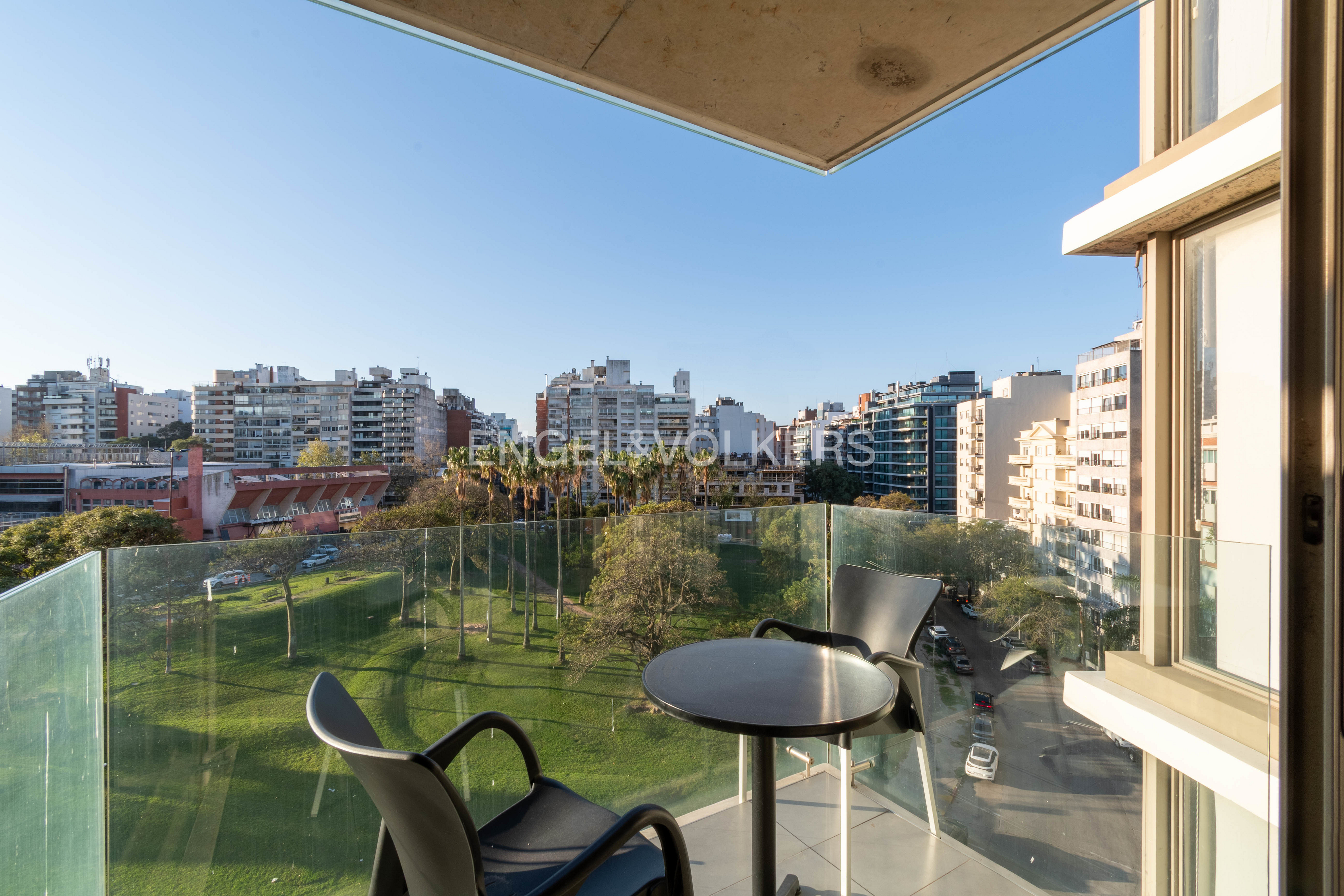 Balcony view with two black chairs and a table overlooking a green park and city buildings under a blue sky.