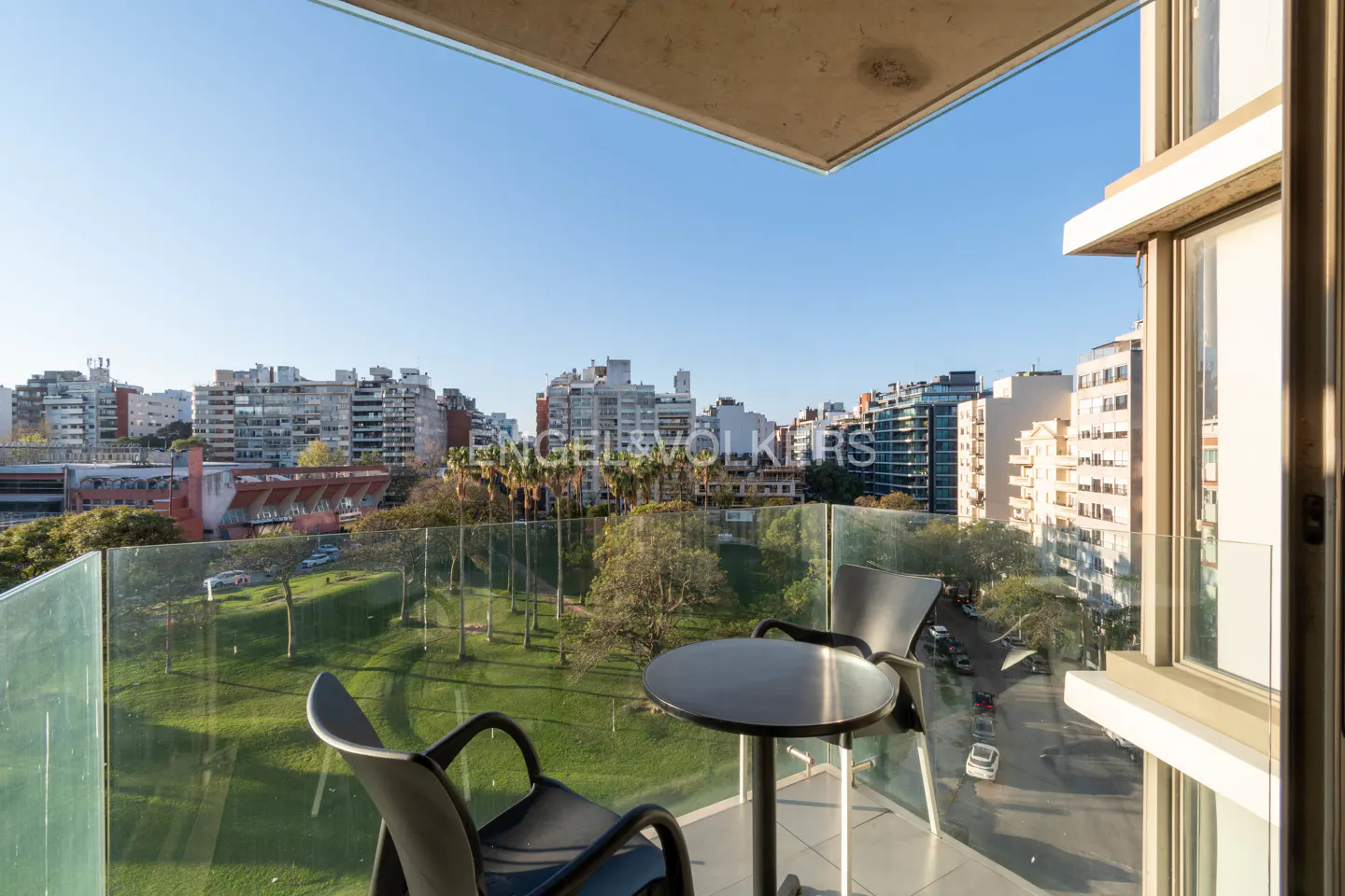 Balcony view with two black chairs and a table overlooking a green park and city buildings under a blue sky.