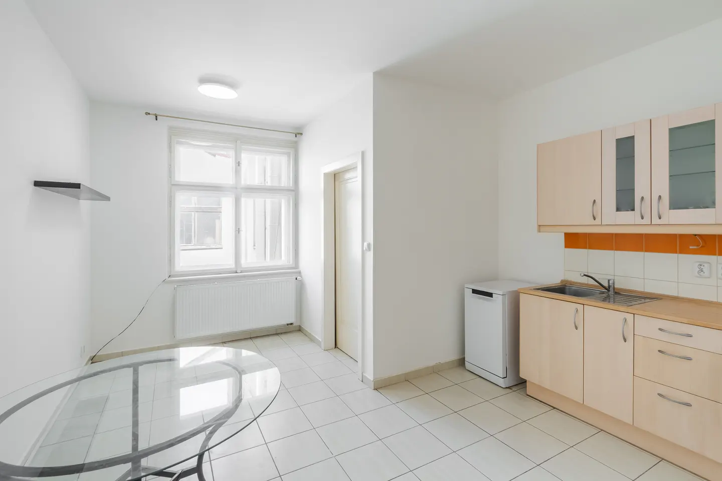 Bright kitchen with white walls, tile floor, and light wood cabinets. A glass table sits near a window with white curtains.