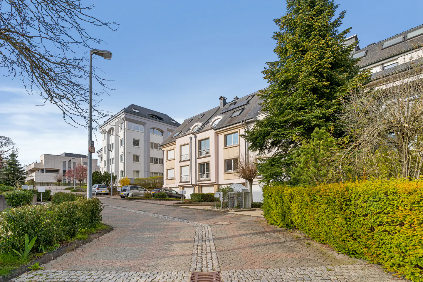 A paved road leads to modern apartments with parked cars, green hedges, and trees under a blue sky.