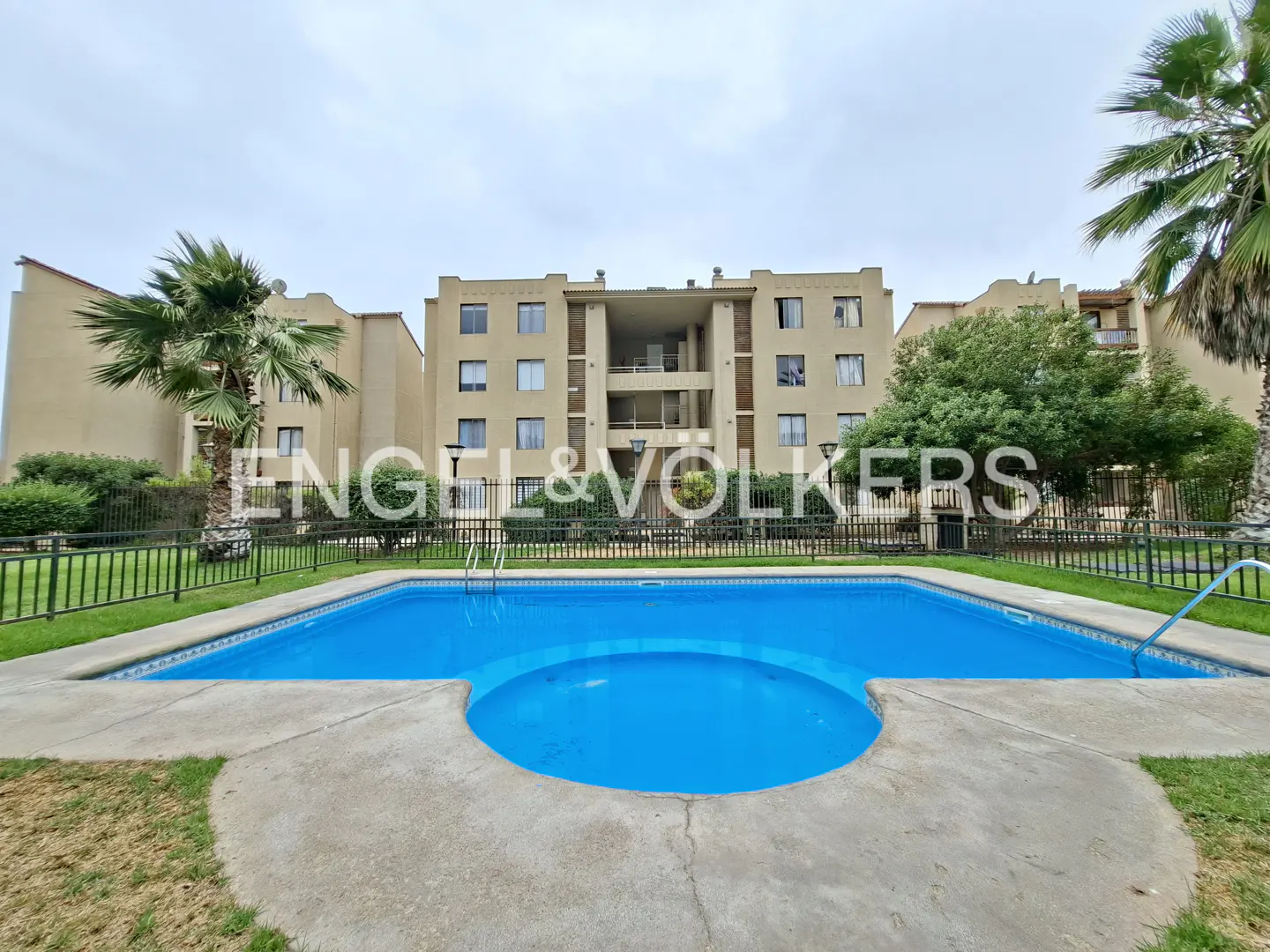 Exterior view of a bright blue swimming pool in front of a tan apartment building with palm trees.