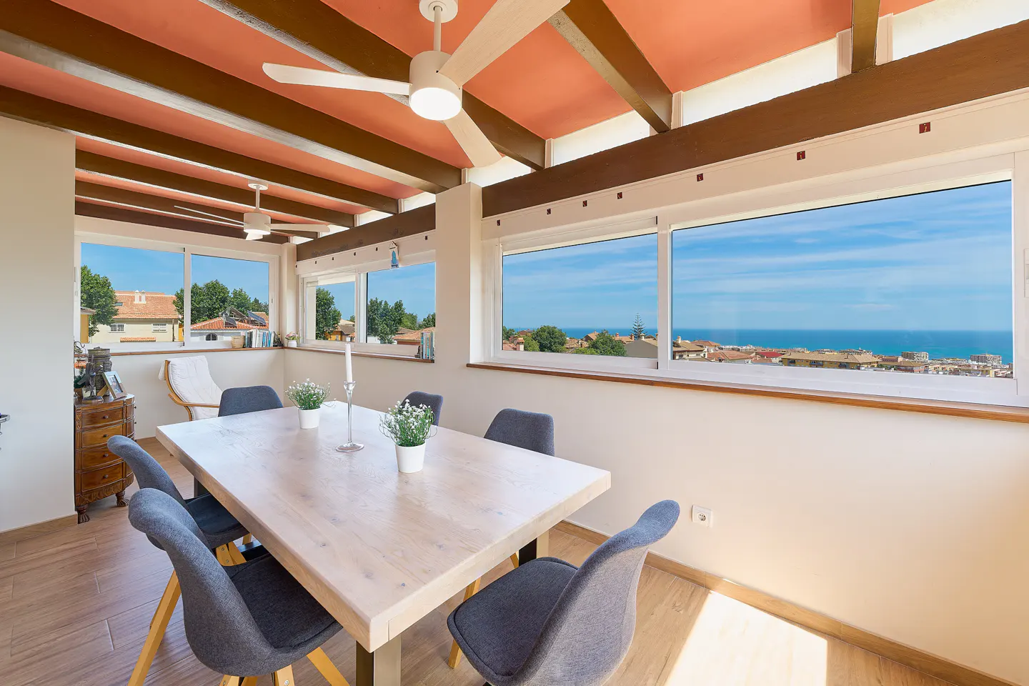 Bright dining room with a light wood table, gray chairs, and large windows overlooking a coastal town and the ocean. Ceiling has exposed beams and a fan.