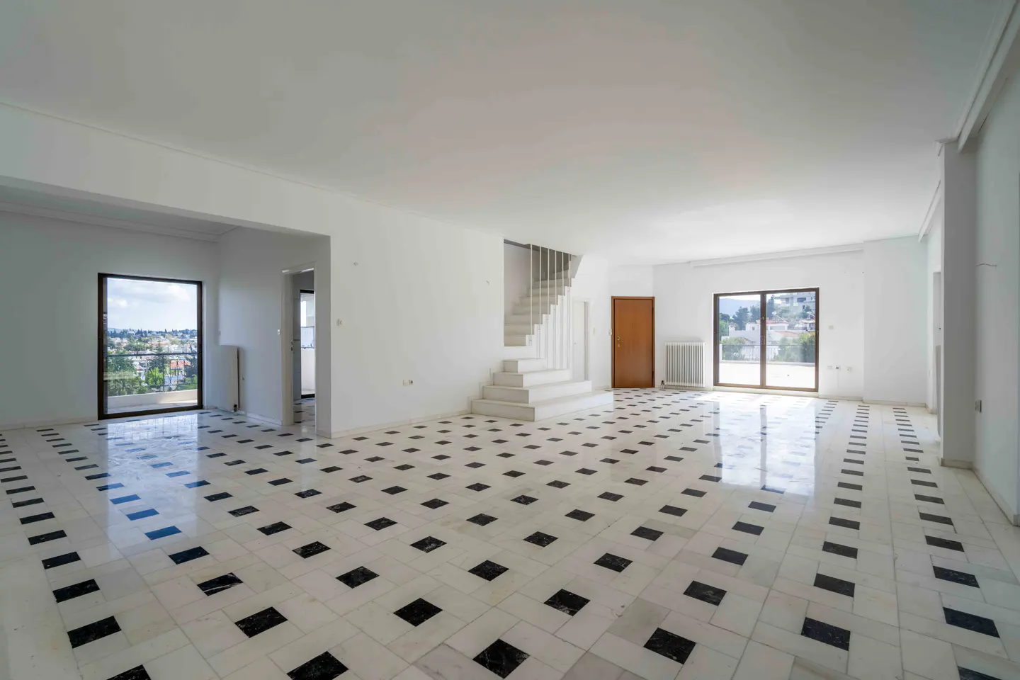 Bright, empty room with white walls, black and white tiled floor, white staircase, and large windows with city views.