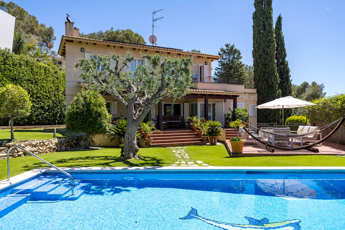 A tan two-story house with a red tile roof overlooks a blue swimming pool with a mosaic dolphin. A hammock and patio furniture sit on a brick patio.