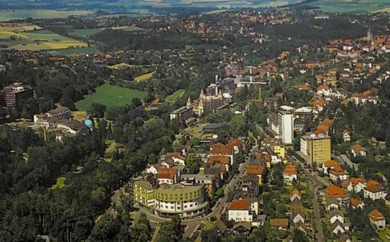 Aerial view of a town nestled among green trees, with buildings of various sizes and colors. Fields and hills are visible in the background.
