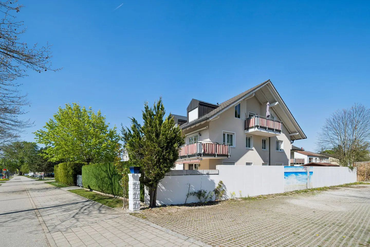 A two-story house with balconies, a white fence, and green trees under a clear blue sky.