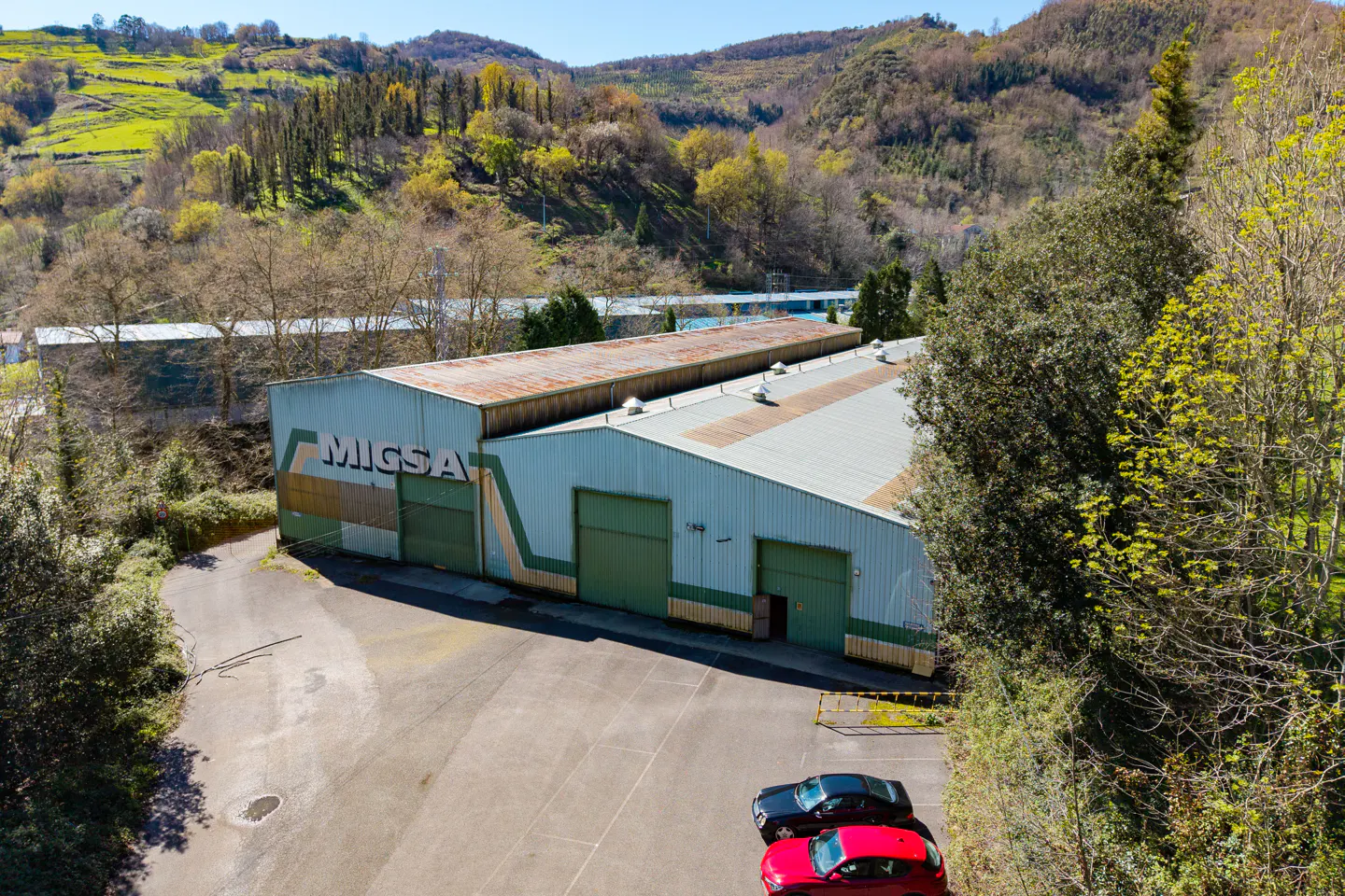 Aerial view of a light blue warehouse with green doors and "MIGSA" on the side, with two cars parked in front. Trees and hills in the background.