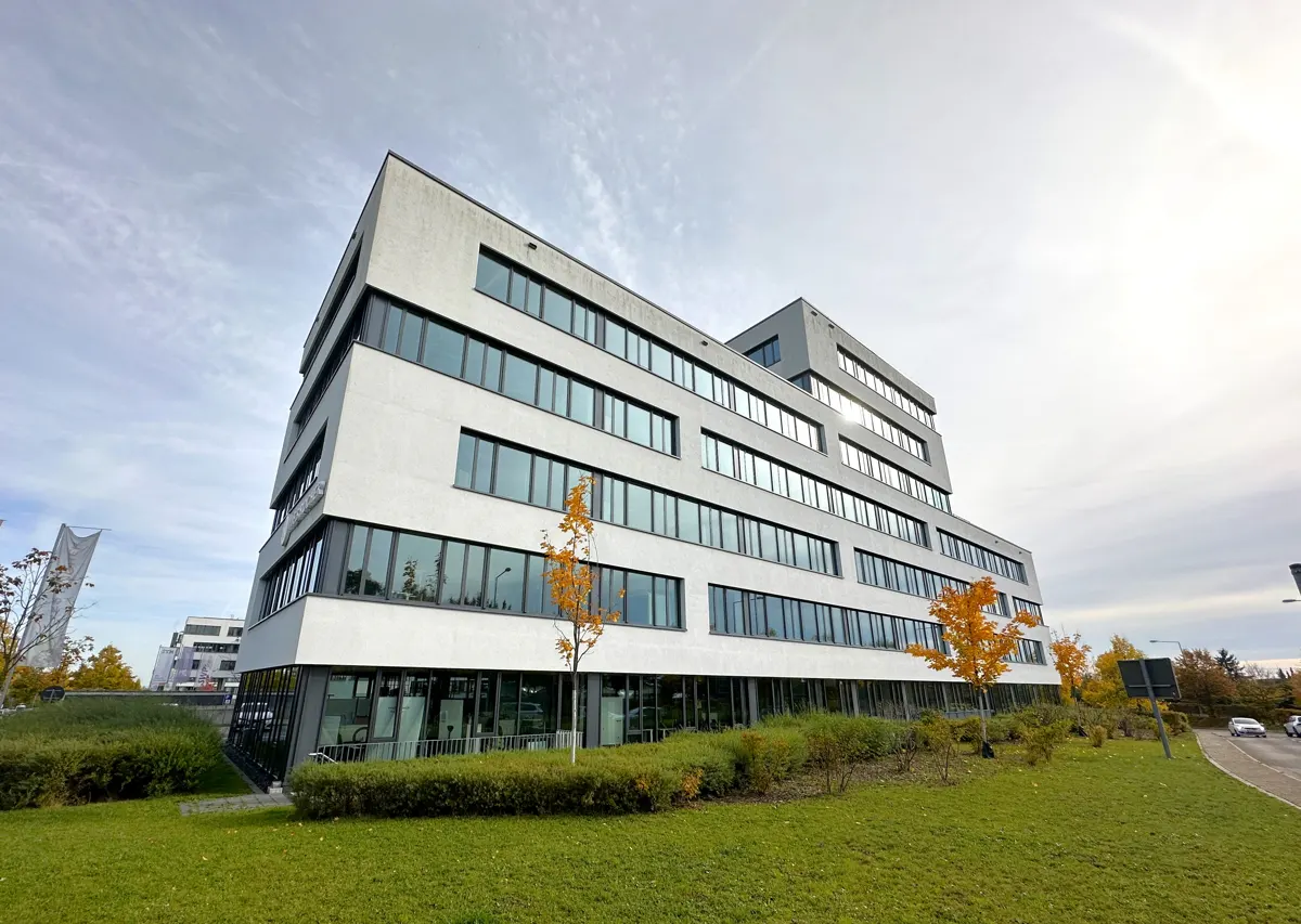 Modern, multi-story office building with white facade and dark-framed windows, surrounded by green lawn and autumn trees.