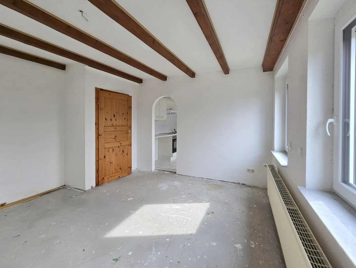Bright, empty room with white walls, exposed wood beams, and a wood door. A glimpse of a white kitchen is visible through an arched doorway.