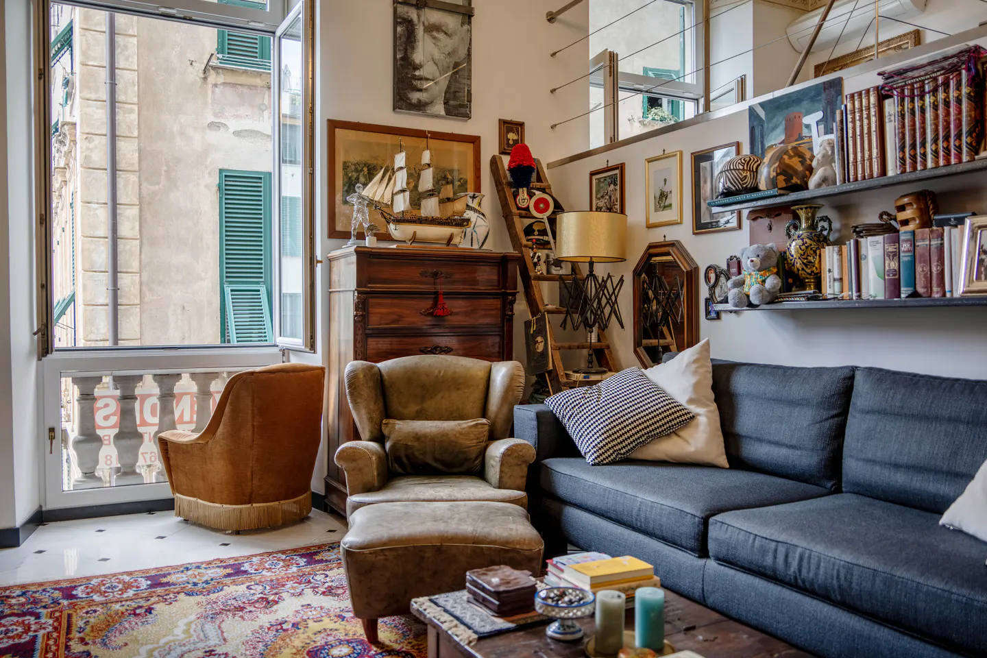 Cozy living room with a blue sofa, brown armchair, and eclectic decor. A window with green shutters overlooks a building. Shelves display books and art.